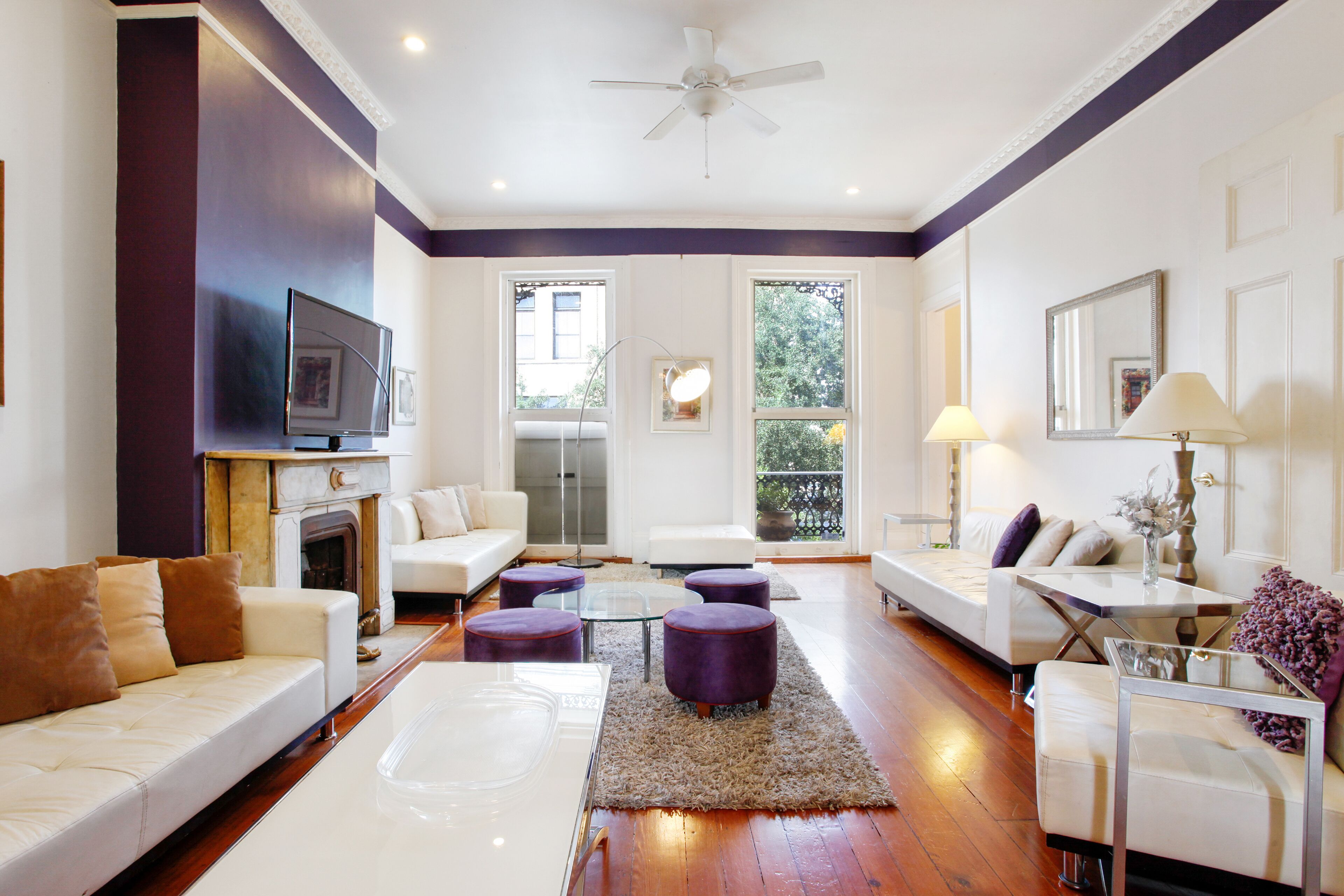 Un salon moderne avec une cheminée, des canapés blancs, des poufs violets et une table basse en verre.