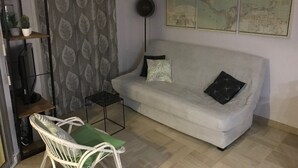 Un salon avec un canapé blanc, deux coussins, une petite table, une chaise, une carte du monde accrochée au mur et un rideau orné d’un motif de feuilles.