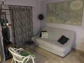 Een woonkamer met een witte bank, twee kussens, een kleine tafel, een stoel, een wereldbol aan de muur en een gordijn met een bladpatroon.