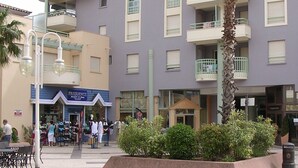 Un immeuble à plusieurs étages doté de balcons, une cour intérieure aménagée avec des sièges et une devanture de magasin appelée « FRÉQUENCE ».