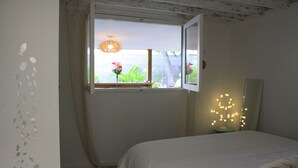A bedroom with a bed, a window with curtains, a wall with a decorative light, and a view of greenery outside. - Jolie CASE CREOLE, LAGON à pied, JARDIN (La Saline-Les-Bains)