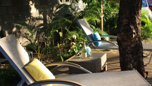 Two lounge chairs with cushions, a bottle, and a glass on a wooden deck surrounded by greenery. - Jolie CASE CREOLE, LAGON à pied, JARDIN (La Saline-Les-Bains)