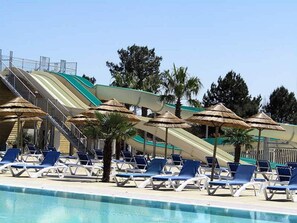 Ein Swimmingpool mit Liegestühlen und Sonnenschirmen, eine Wasserrutsche und im Hintergrund Palmen.