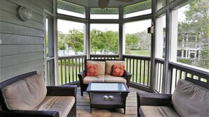 Ein überdachter Balkon mit Deckenventilator, einer gepolsterten Couch mit roten und beigen Kissen sowie einem Glastisch.