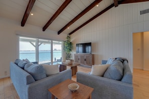 A living room with two grey sofas, a wooden coffee table, a TV on a wooden cabinet, and a large window with a view of the sea.