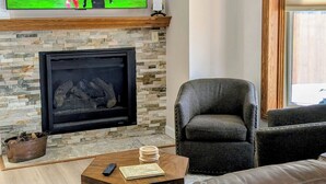 Un salon avec une cheminée en pierre, un canapé en cuir, une table basse avec une télécommande et un téléviseur diffusant un match de football.
