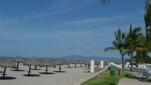 A beach with thatched umbrellas, lounge chairs, and palm trees. - Luxurious newly renovated BeachFront Condo & Vidanta Membership (Nuevo Vallarta)