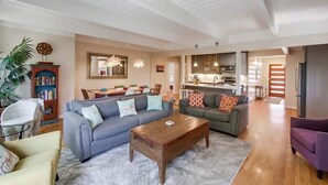 Un vaste salon comprenant un grand canapé, une table basse et un coin repas avec des chaises et une table. On aperçoit une cuisine au fond.