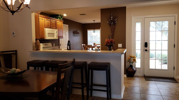 A kitchen with wooden cabinets, a white microwave, and a dining area with a table and chairs. - Walk to Lake Sumter Landing! Golf cart included! (The Villages)
