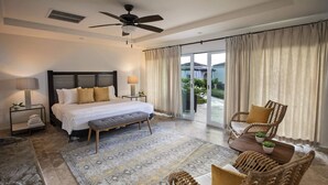 A spacious bedroom with a large bed, a ceiling fan, a bedside table with a lamp, a bench, and a view of the outdoor area through a sliding glass door. - Bougainvillea Large 3 Bedroom with Bonus Room (St. John)