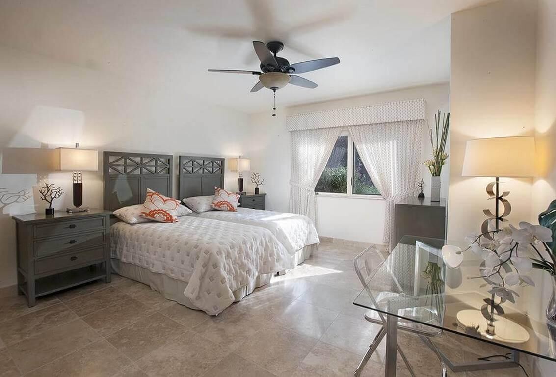 A bedroom with a bed, bedside tables, a ceiling fan, a window with curtains, and a glass table with a vase.