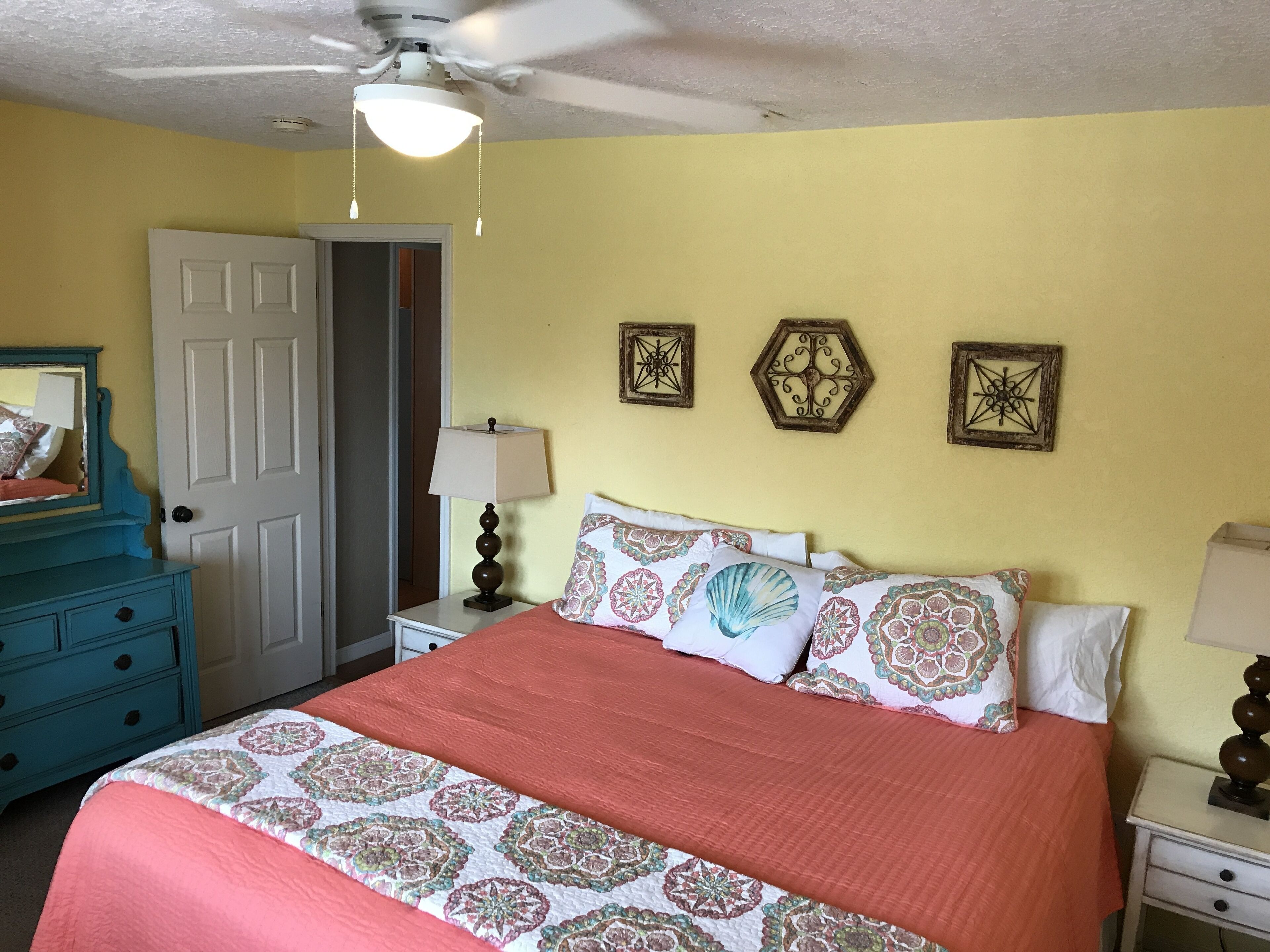 Une chambre à coucher comprenant un lit, une commode, une table de chevet, un ventilateur de plafond et des œuvres d’art décoratives accrochées au mur.