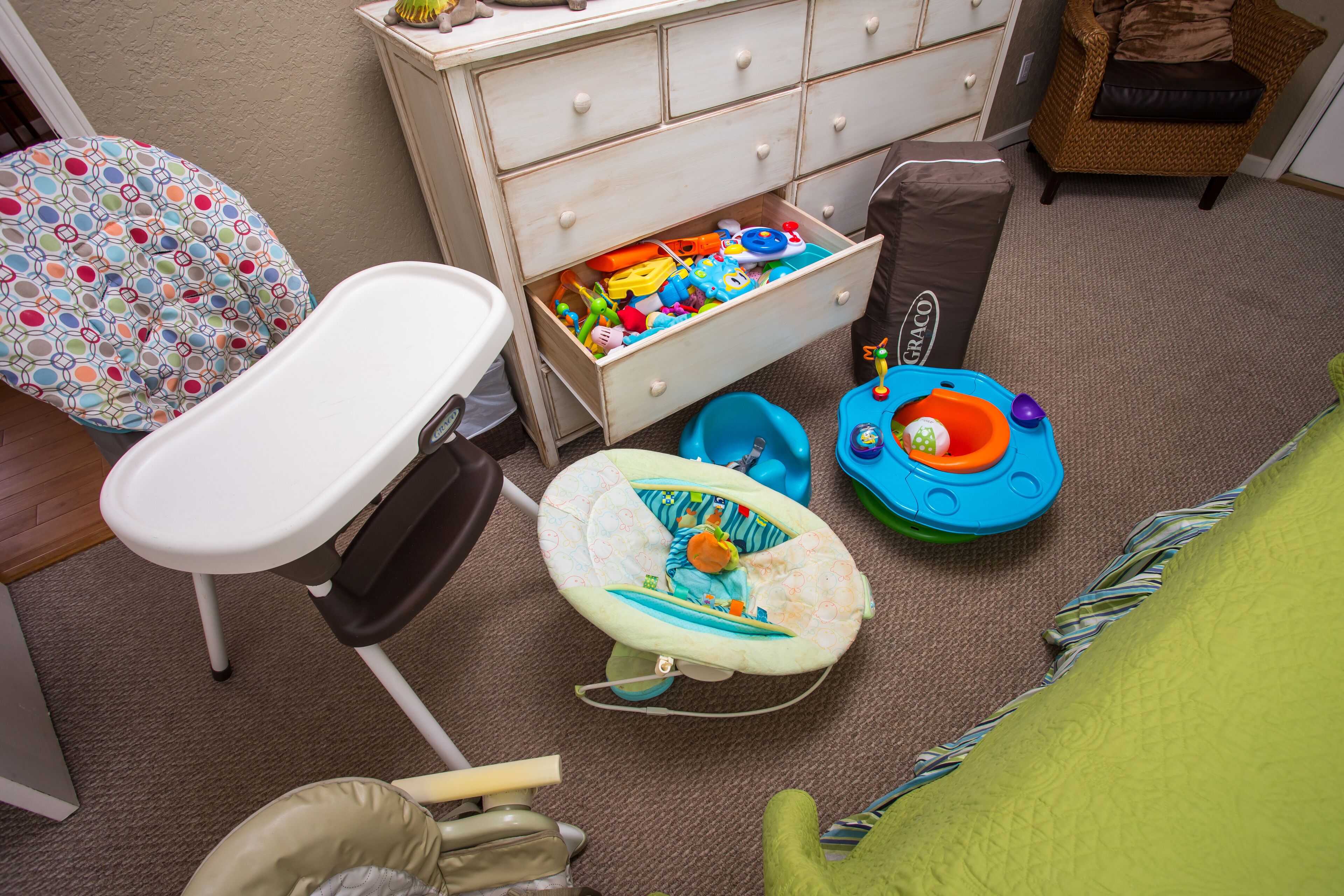 A child's play area with a high chair, a baby rocker, and a storage chest filled with toys.