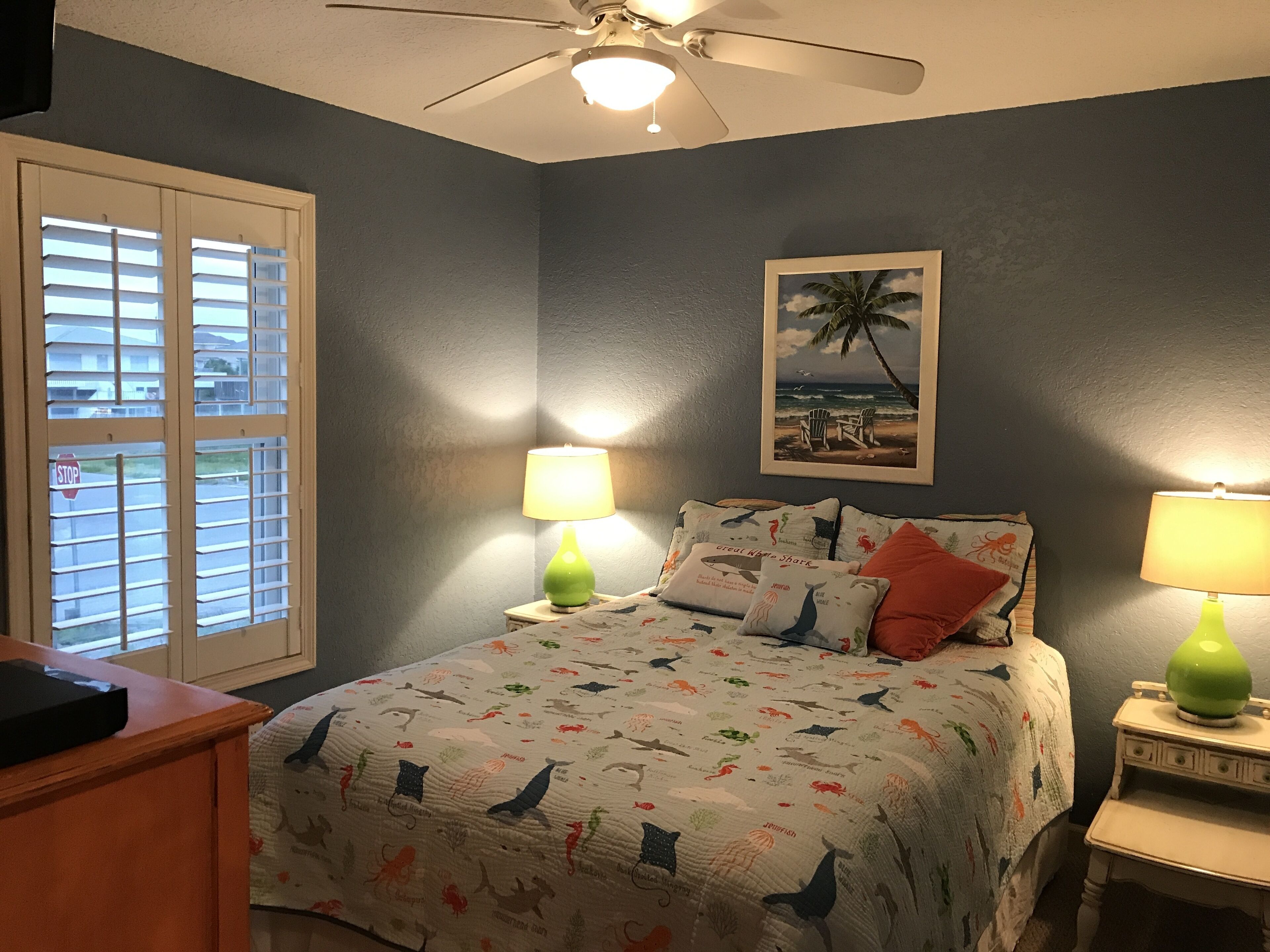 A bedroom with a bed, two bedside lamps, a ceiling fan, a window with blinds, and a framed picture on the wall.