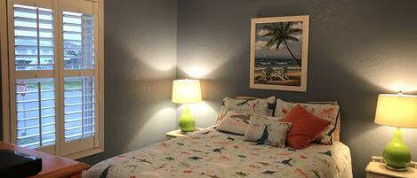 A bedroom with a bed, two bedside lamps, a ceiling fan, a window with blinds, and a framed picture on the wall.