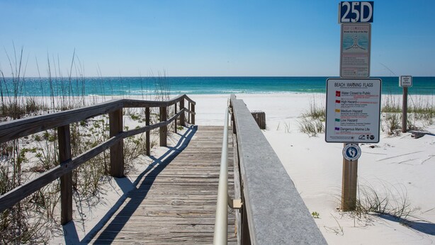 Une promenade en bois mène à une plage sous un ciel dégagé. Un panneau indique les drapeaux de signalisation de la plage et un marqueur de distance de 25 unités.