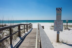 Une promenade en bois mène à une plage sous un ciel dégagé. Un panneau indique les drapeaux de signalisation de la plage et un marqueur de distance de 25 unités.