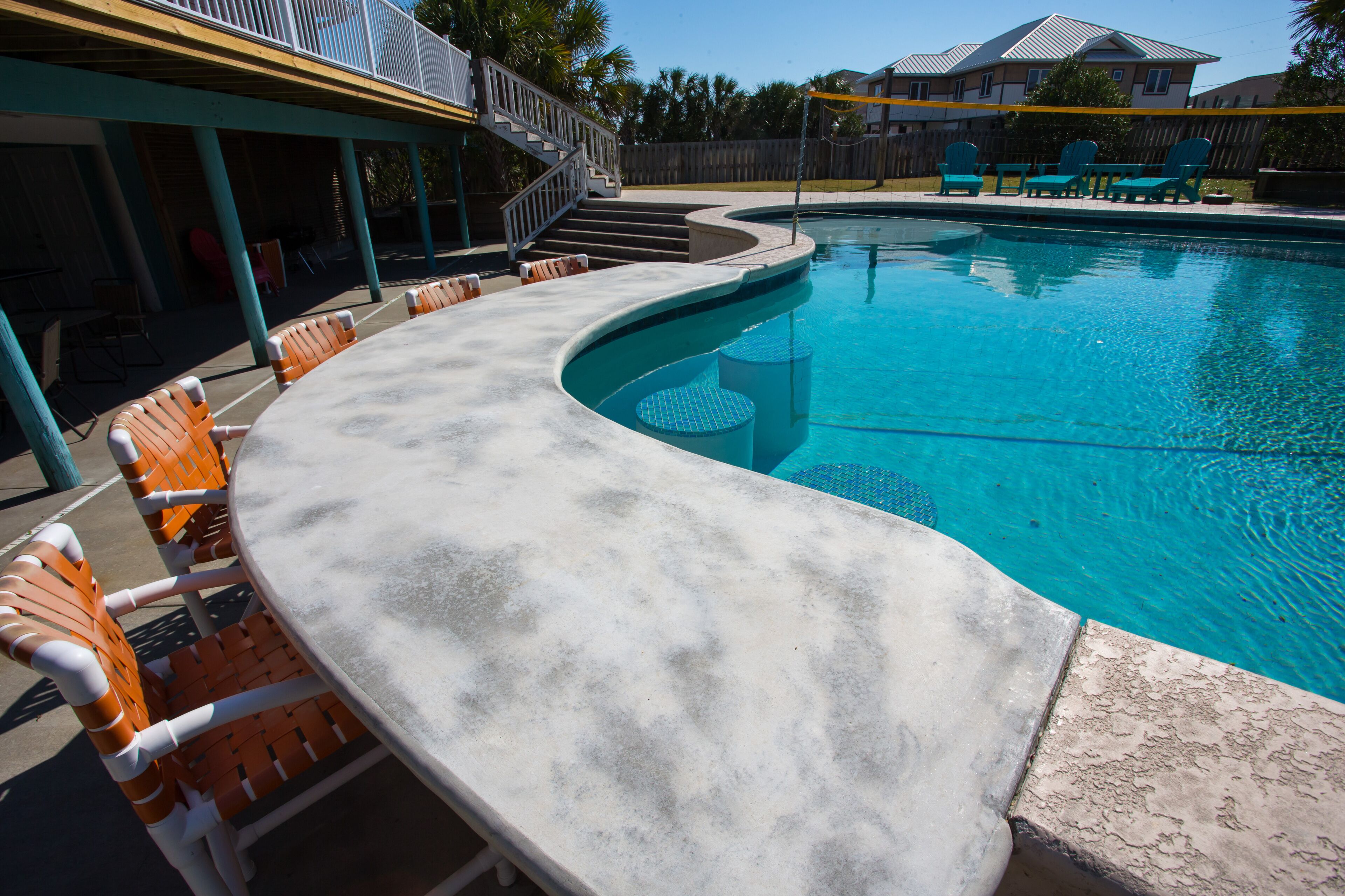 Zona de piscina con un deck curvo de hormigón, sillones de relax naranja y blanco, y un área de estar cubierta con escaleras que conducen a un balcón.