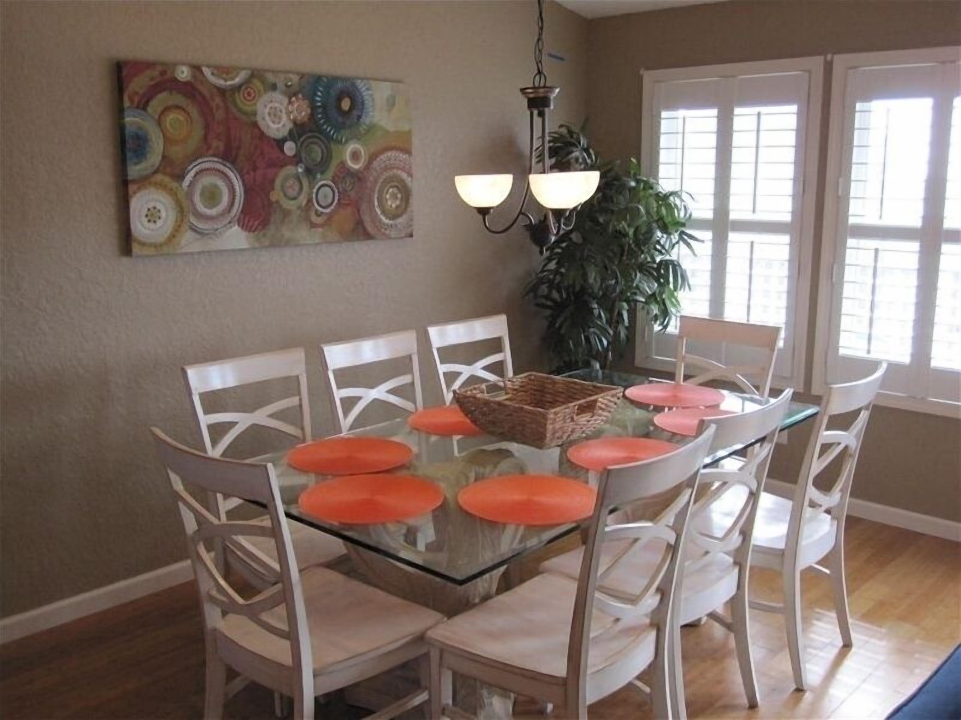 A dining area with a glass table surrounded by white chairs with orange cushions, a hanging light fixture, and a colorful abstract painting on the wall.