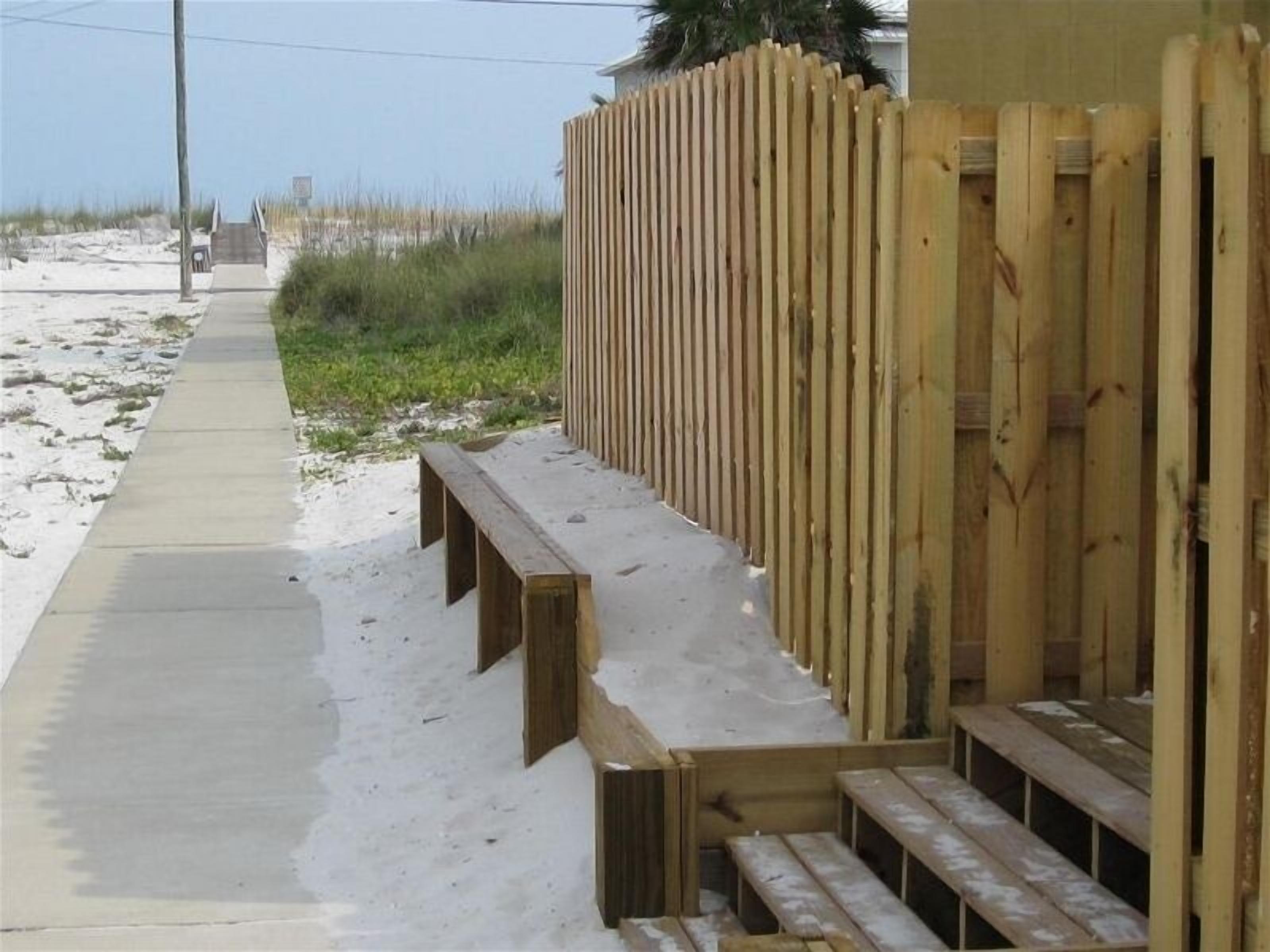 Una terraza de madera con bancos, un camino de hormigón y una playa arenosa al fondo.