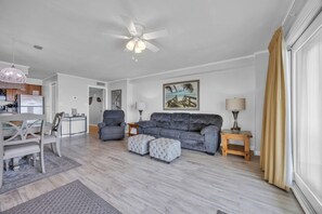 Ein modernes Wohnzimmer mit einem grauen Sofa, zwei Poufs, einem Esstisch mit Stühlen, einem Deckenventilator und einem Gemälde an der Wand.