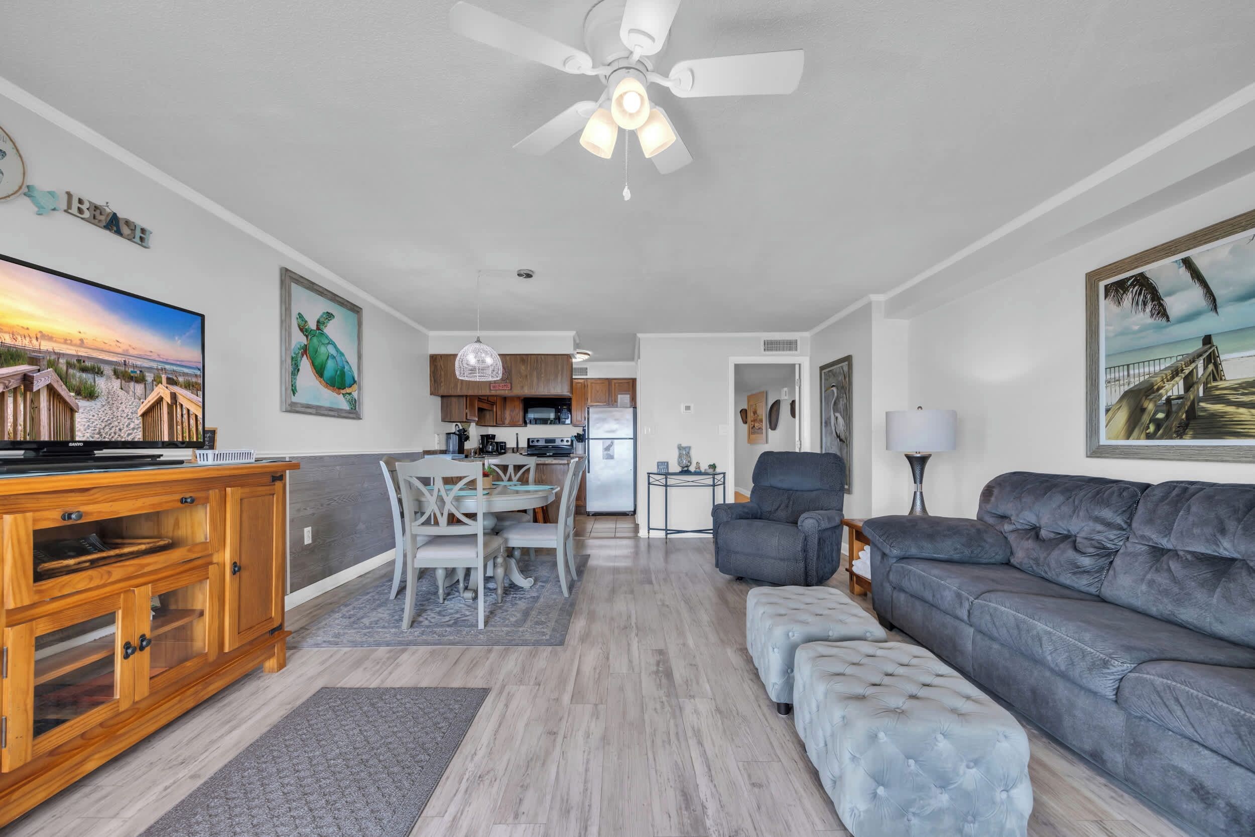 A spacious living room with a grey sofa, a wooden entertainment unit, and a dining area with a white table and chairs. A kitchen with modern appliances is visible in the background.