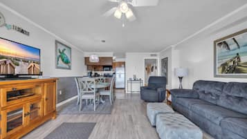 A spacious living room with a grey sofa, a wooden entertainment unit, and a dining area with a white table and chairs. A kitchen with modern appliances is visible in the background.