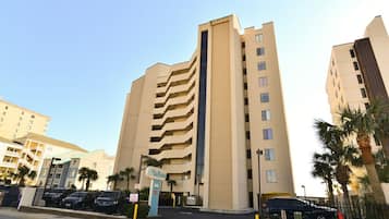 A multi-story hotel with a central vertical feature, a parking area with cars, and a clear sky.