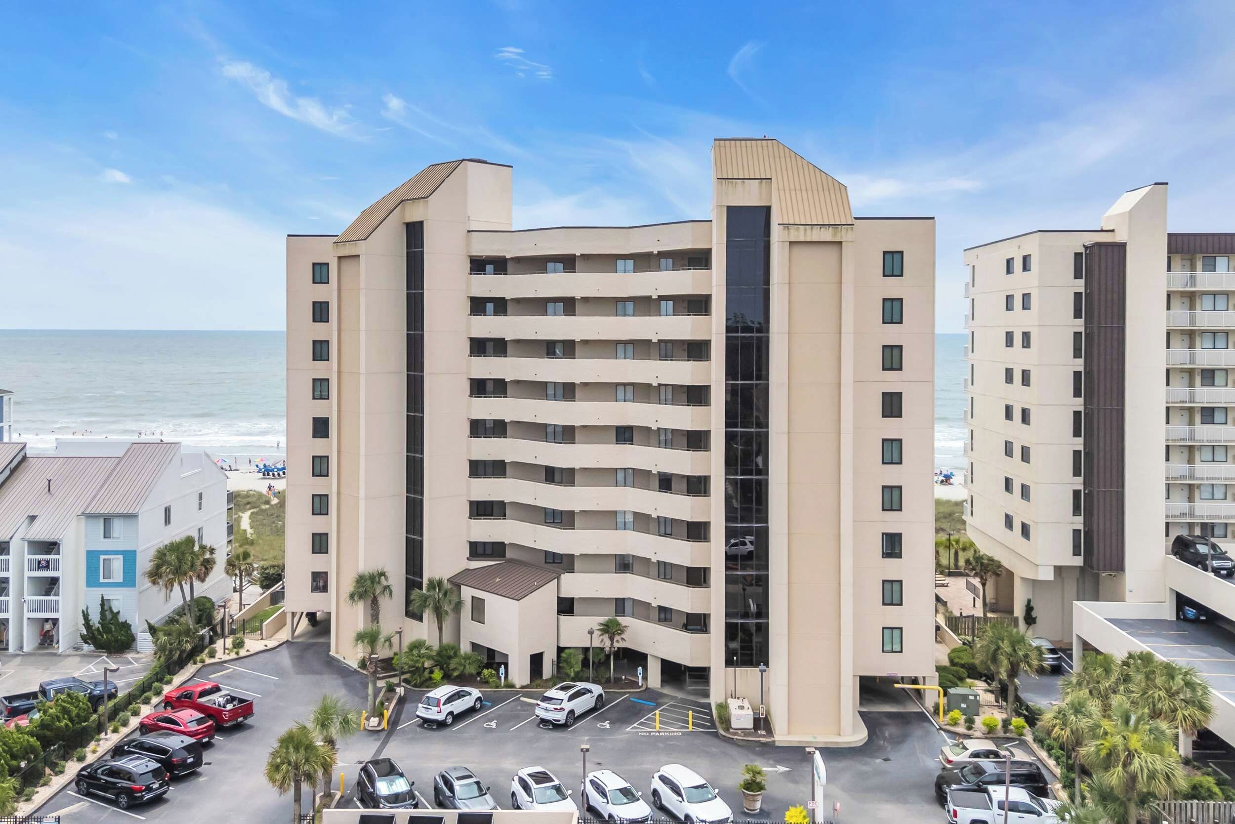 A multi-story hotel with a parking lot in front, a beach view, and a modern architectural design.