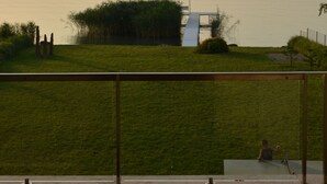Vista de un lago tranquilo con una cadena montañosa distante, un área verde con una pequeña construcción y una persona jugando con un juguete.