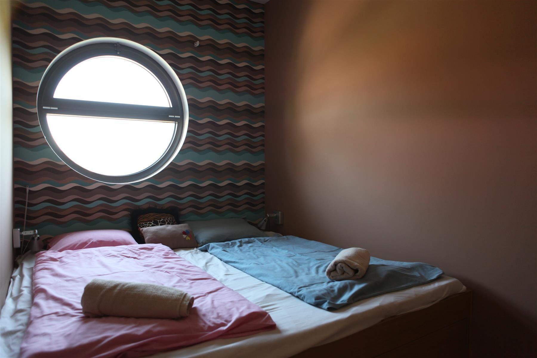 A bedroom with a bed, pillows, a round window, and a patterned wall.