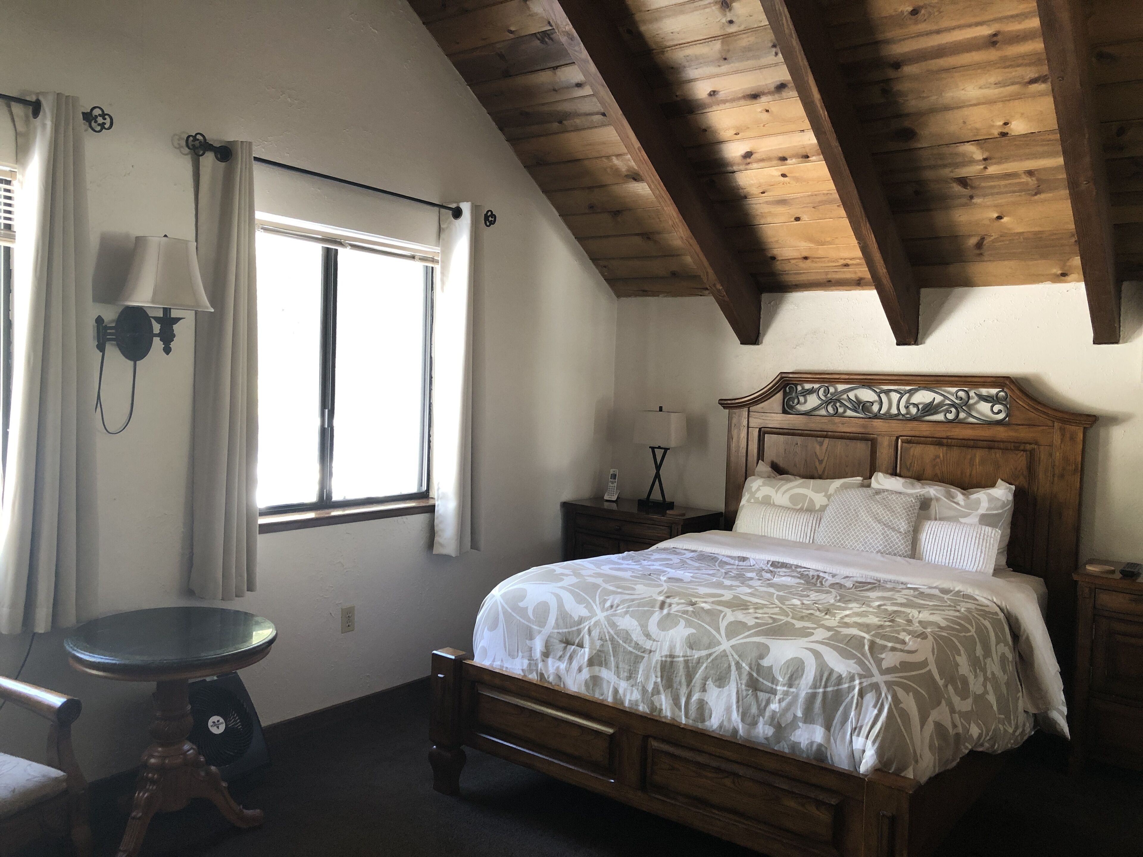 A bedroom with a wooden ceiling, a bed with a patterned bedspread, a nightstand with a lamp, and a window with curtains.