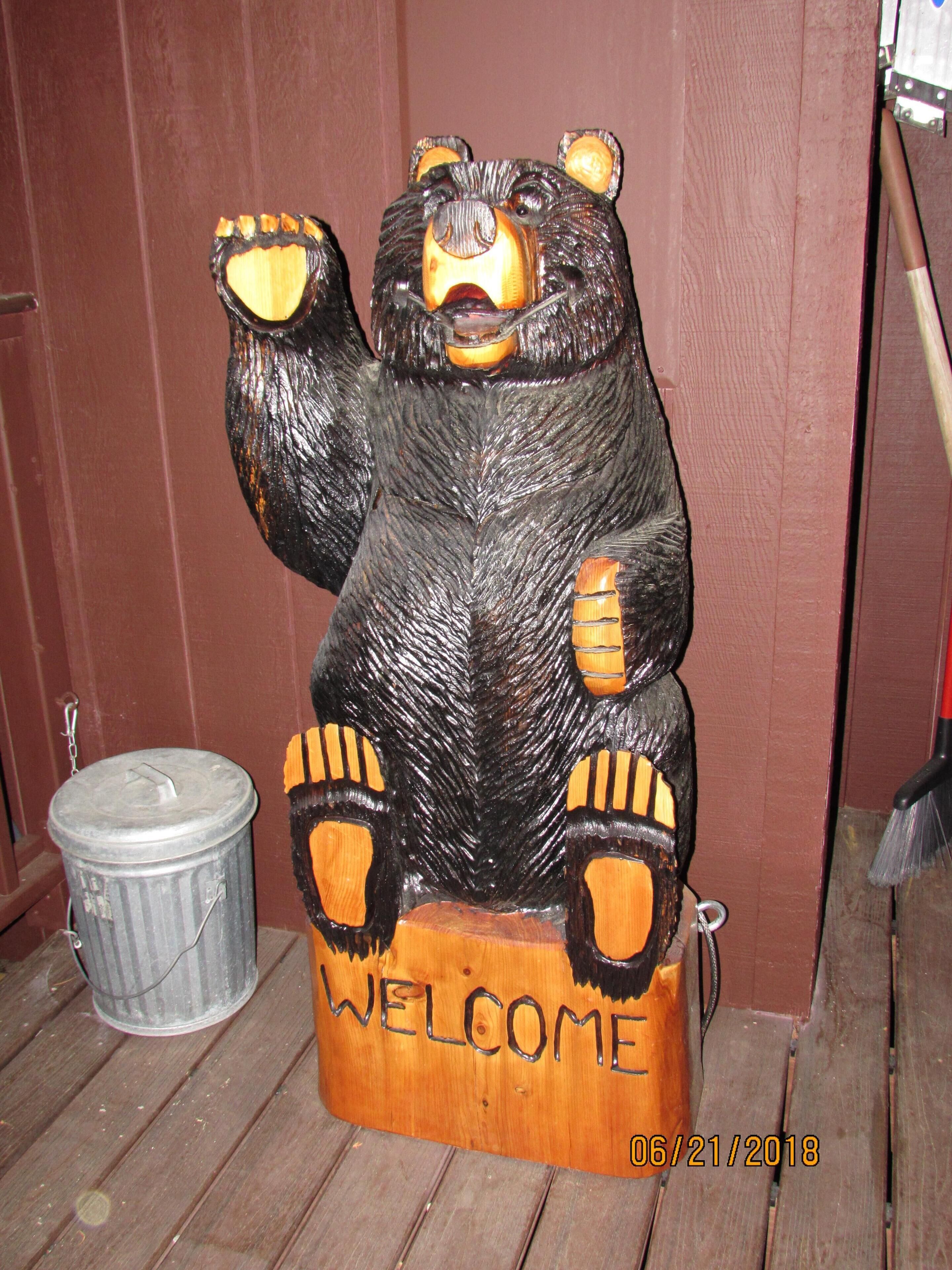 A wooden bear sculpture with a 'WELCOME' sign at its base, positioned outdoors against a wooden wall.