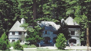 A lakeside property with a large house featuring a stone tower and a smaller building with a pointed roof.
