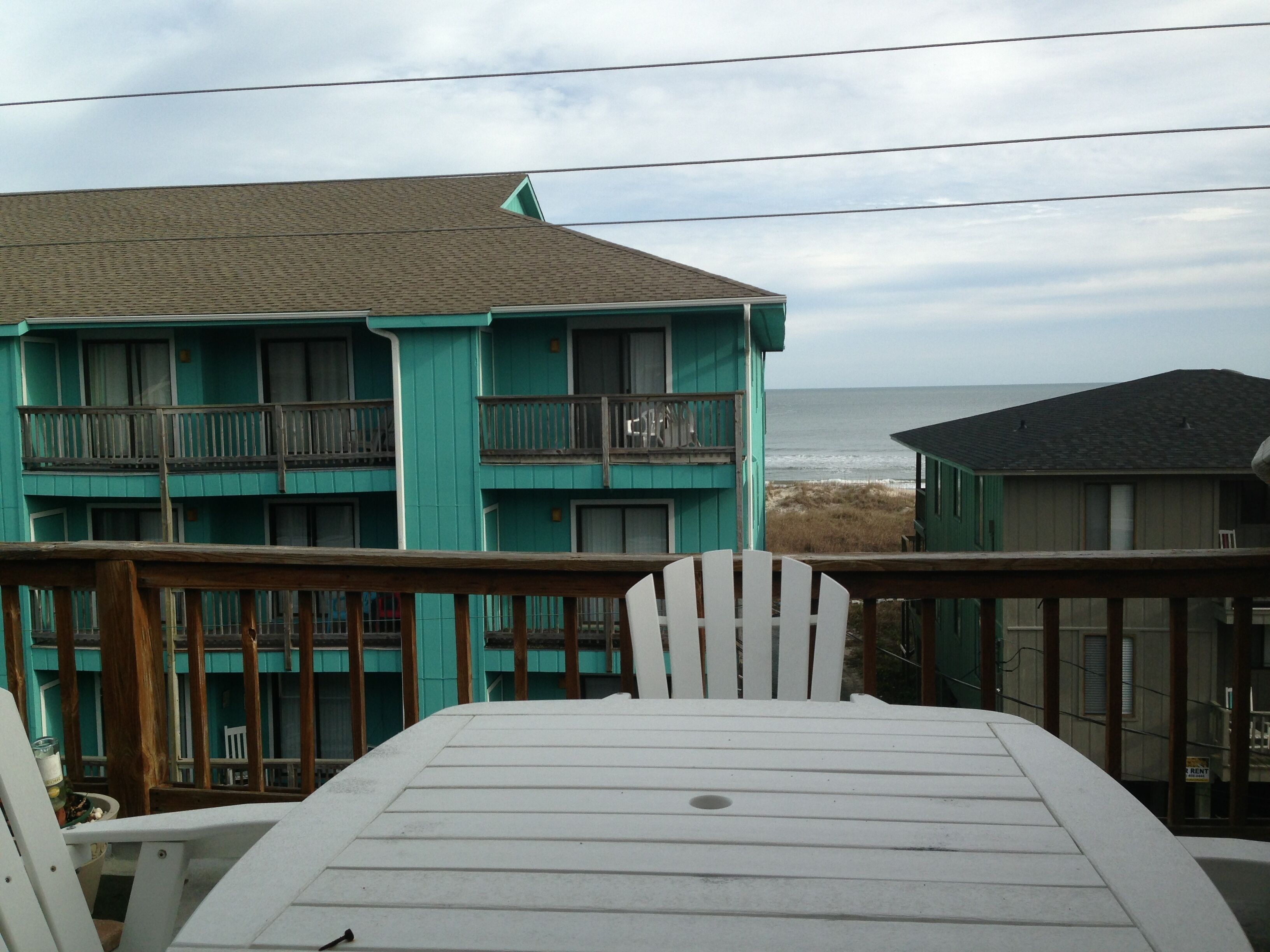 A turquoise building with balconies, a beach view, and a wooden deck with a table and chairs.