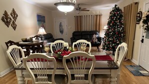 Comedor con mesa de madera y sillas blancas, un árbol de Navidad y un sofá de cuero negro al fondo.