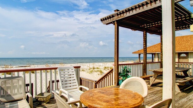 Una terraza de madera con mobiliario exterior, incluyendo sillas y una mesa, con vistas a la playa.