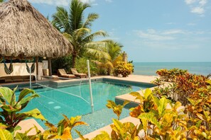 Zona de piscina amb gandules, una construcció amb teulada de palla i plantes tropicals.
