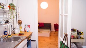 Une cuisine équipée d’un évier, d’étagères et d’une plante suspendue. On aperçoit, à travers une porte ouverte, un coin repas avec une table et des chaises.