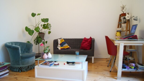 Un salon avec un fauteuil bleu, une table basse en verre, un canapé gris agrémenté de coussins rouges et jaunes, une plante en pot et une petite bibliothèque.