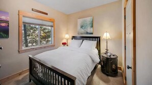 A bedroom with a bed, bedside lamps, a window with a view of trees, and a painting on the wall. - Arrowhead Properties Unit 4 (West Yellowstone)
