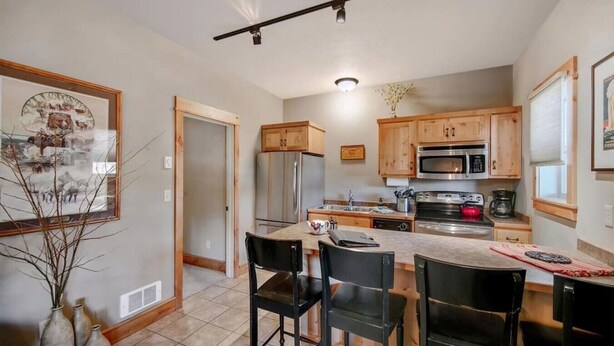 A kitchen with wooden cabinets, a stainless steel refrigerator, a microwave, and a stove. There are two black chairs and a small table with a vase. - Arrowhead Properties Unit 4 (West Yellowstone)