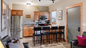 A compact kitchen with wooden cabinets, a refrigerator, a microwave, and a dining area with four chairs. - Arrowhead Properties Unit 4 (West Yellowstone)