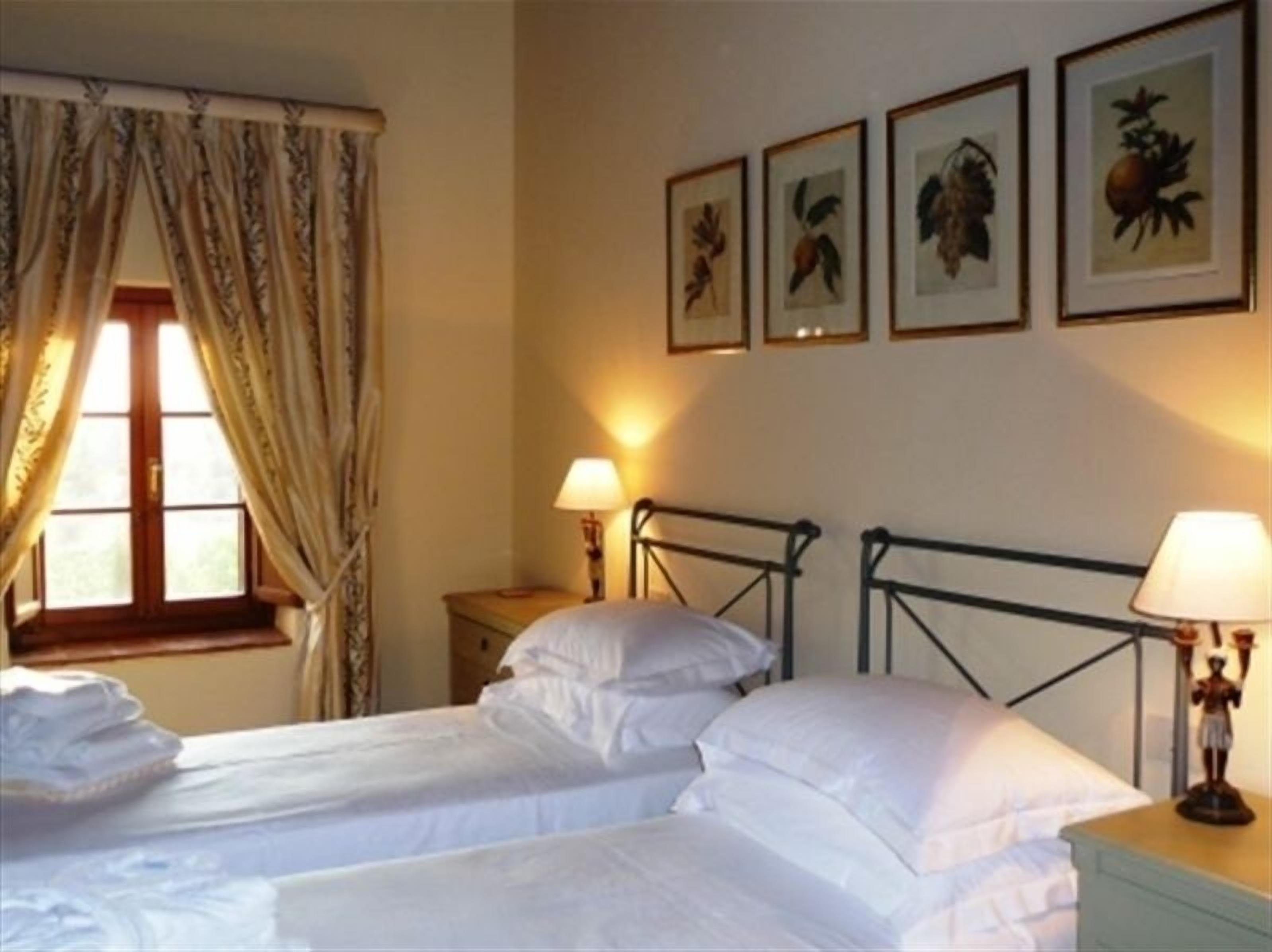 Une chambre avec deux lits, une fenêtre avec des rideaux, une table de chevet avec une lampe et, au mur, des reproductions encadrées de motifs botaniques.