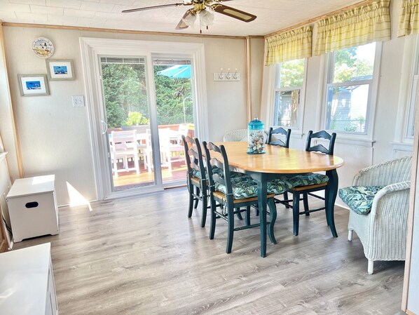 Un comedor con una mesa redonda de madera, sillas tapizadas en verde oscuro y una silla de mimbre con cojín estampado. Se observa un ventilador de techo, una ventana con un cortinero amarillo y vistas a una terraza con mobiliario exterior.