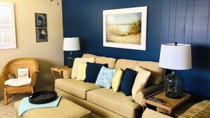 Una sala de estar con un sofá color beige, dos mesitas auxiliares, un sillón de mimbre y un cuadro con una imagen de playa en la pared.