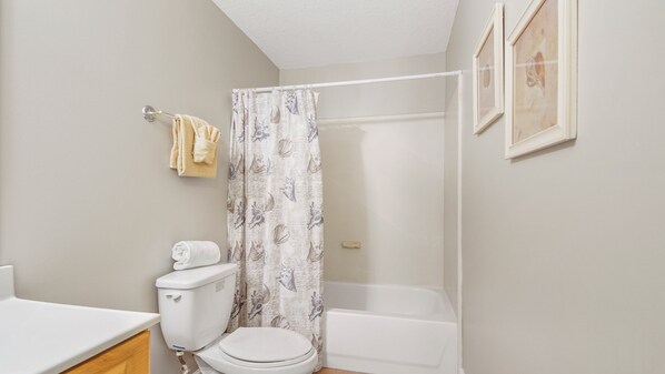 A bathroom with a toilet, a bathtub, and a shower curtain with a shell pattern. There are towels hanging on a rack and a framed picture on the wall. - Emerald Isle 1701 - BEACH CHAIRS INCLUDED! Affordable rates! (Panama City Beach)