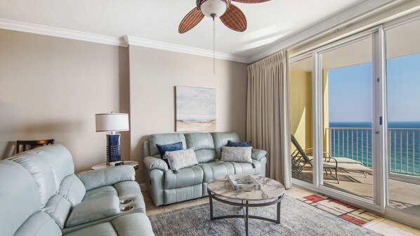 A modern living room with a sofa set, a coffee table, and a view of the ocean through large sliding doors. - Emerald Isle 1701 - BEACH CHAIRS INCLUDED! Affordable rates! (Panama City Beach)