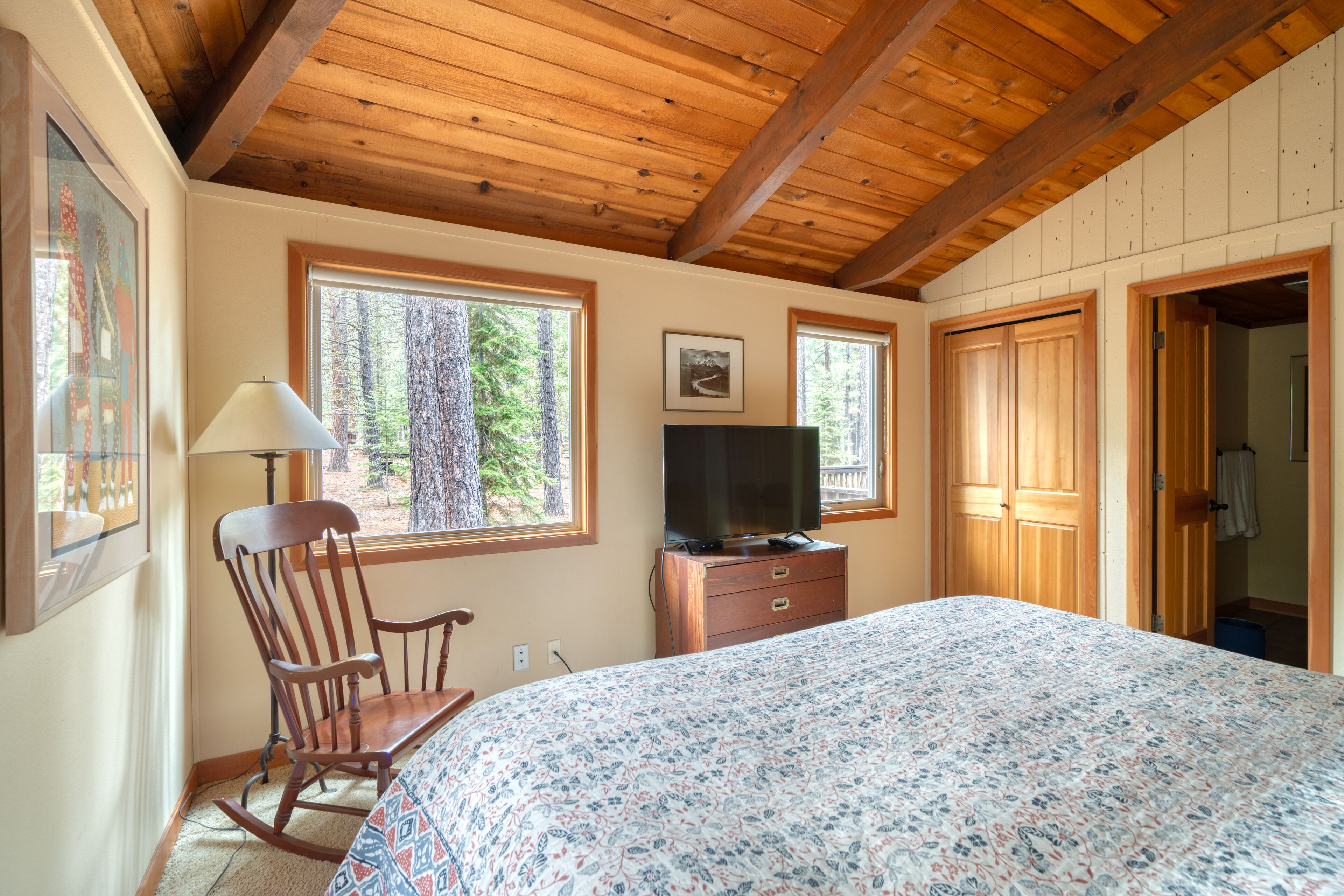 A bedroom with a wooden ceiling, a bed with a floral bedspread, a rocking chair, a dresser, a television, and a window with a view of trees.