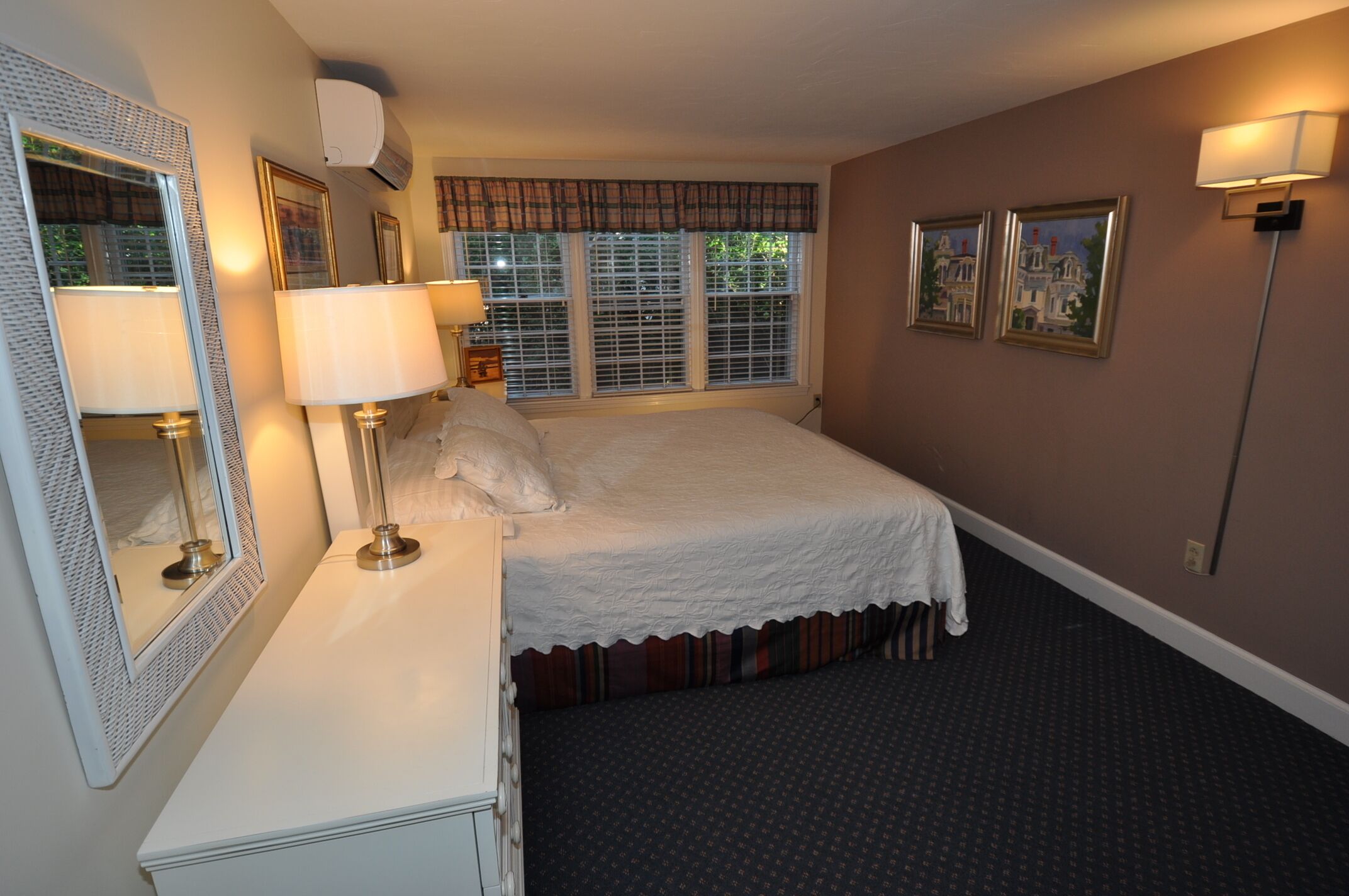 A hotel room with a bed, a dresser, a mirror, two wall-mounted lamps, and a window with blinds.